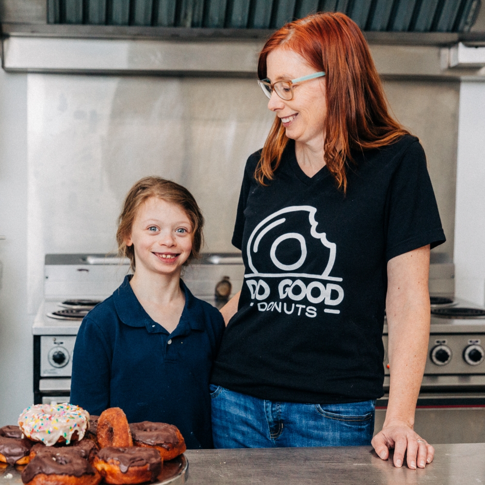 Melanie Côté, Owner, Do Good Donuts. Melanie is at her bakery shop, smiling, standing with a child. 