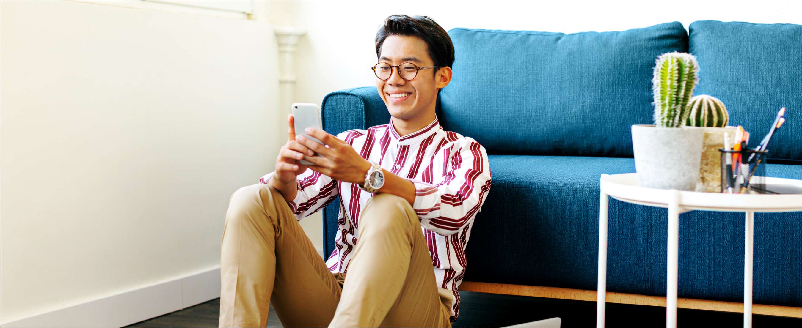 Smiling Asian man looking at smart phone while sitting on carpet against sofa at home