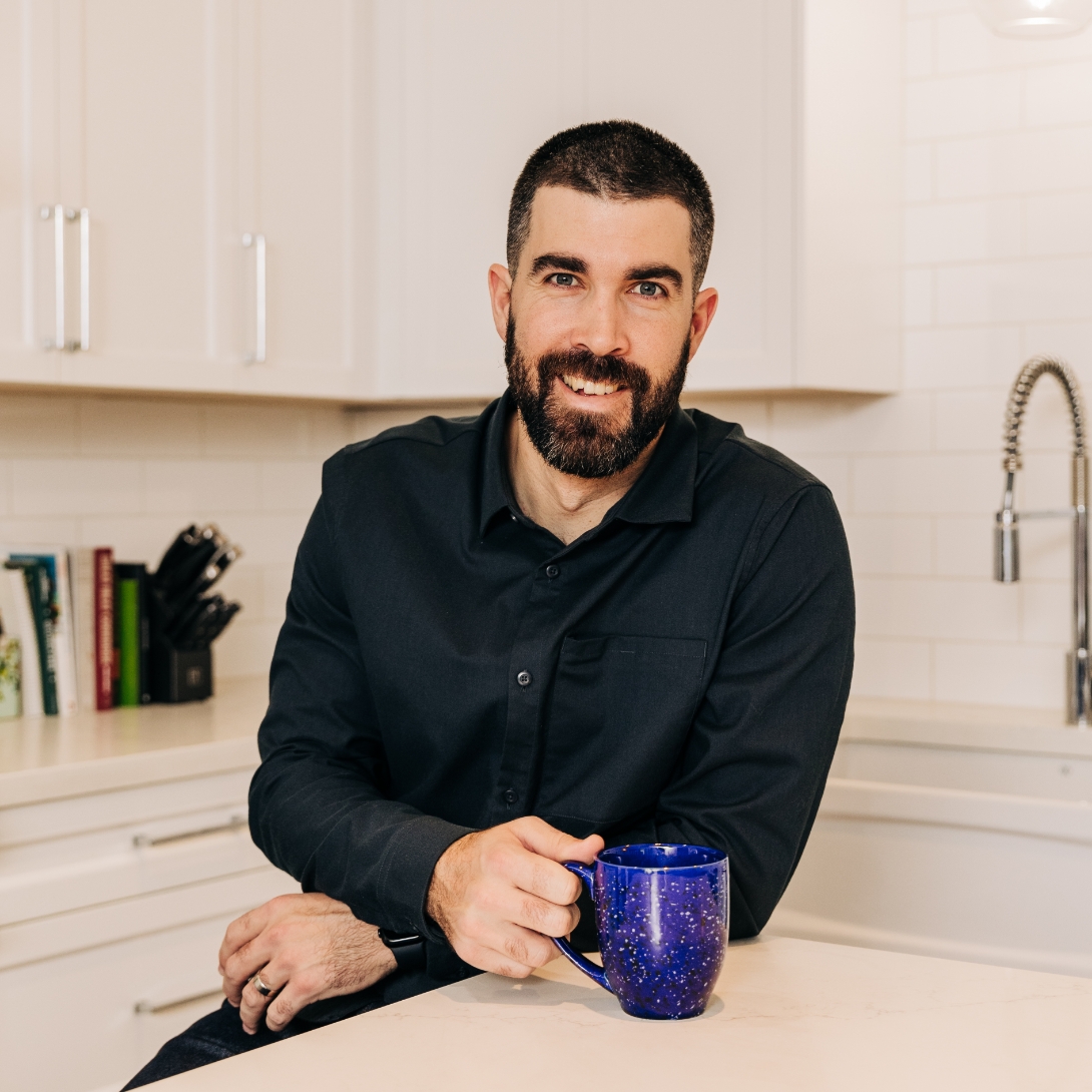A smiling man sits at a kitchen table with a cup of coffee.