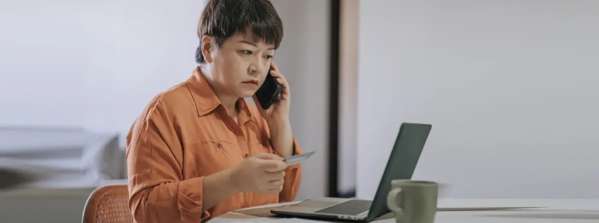 Woman on the phone, holding credit card, looking at computer screen with a worried expression.