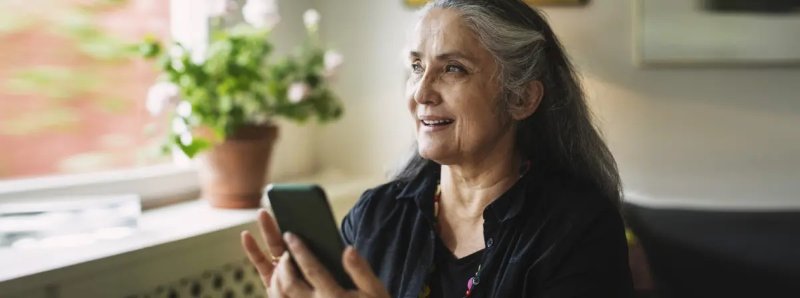 A mature woman with medium skin tone and long, greying hair, holds her phone in front of her, smiling slightly as she speaks. With flowers on the windowsill beside her, she looks out the window with an expression of familiarity, as if she recognizes the voice on the phone.