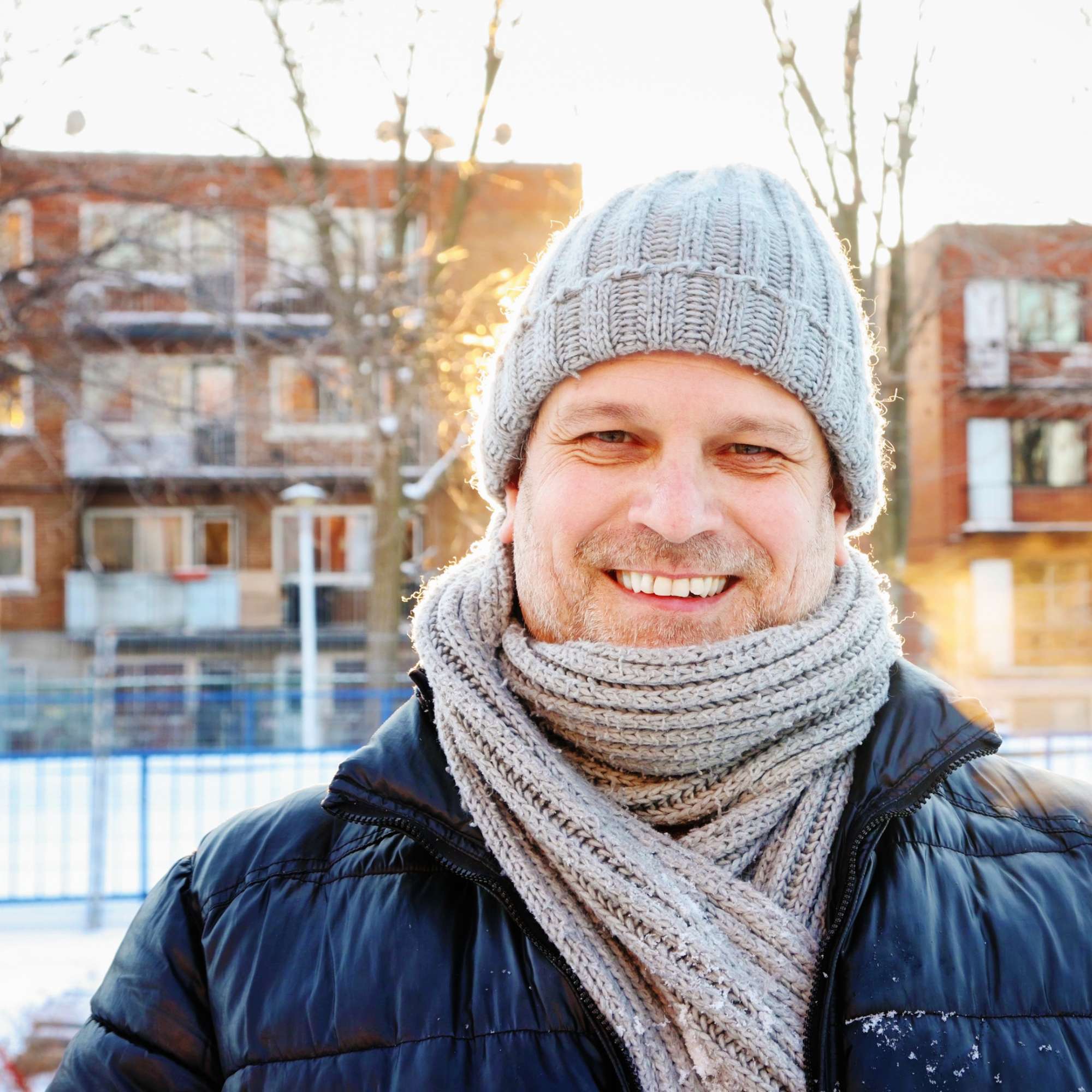 A smiling man stands in a snowy city park, bundled up. He has light skin, a blonde beard, and is wearing a grey hat.