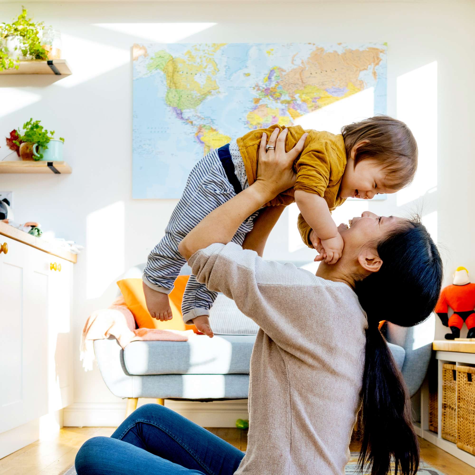 A woman with light skin tone and long black hair is sitting on the floor of a sunny living room, smiling as she lifts her laughing baby up in the air. A woman with light skin tone and long black hair is sitting on the floor of a sunny living room, smiling as she lifts her laughing baby up in the air. 