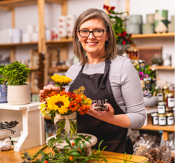 Woman smiling in her flower shop as she prepares a flower arrangement. She has medium brown short hair and glasses.