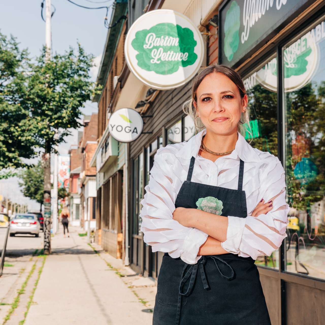 A woman wearing a white shirt and a black apron stands confidently in front of a store named "Boston Lettuce" on a sidewalk. 