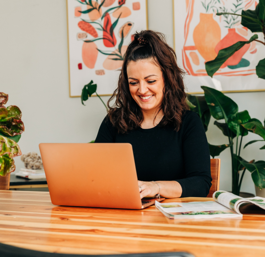 A woman sits at a wooden table, using a laptop, with two colorful botanical posters on the wall behind her and a magazine open beside her.