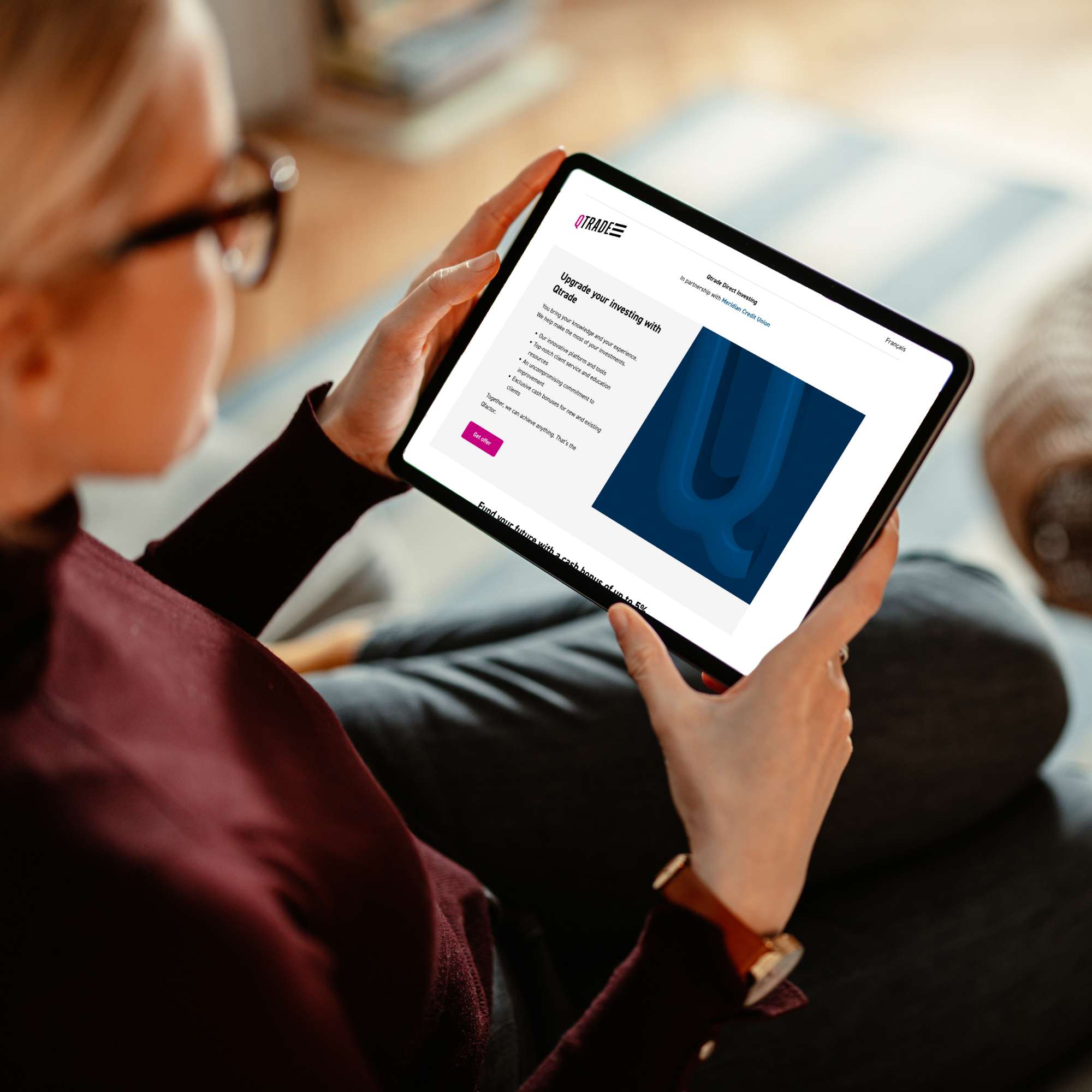 Woman with blonde hair and glasses is holding a tablet. The screen shows the Qtrade website.