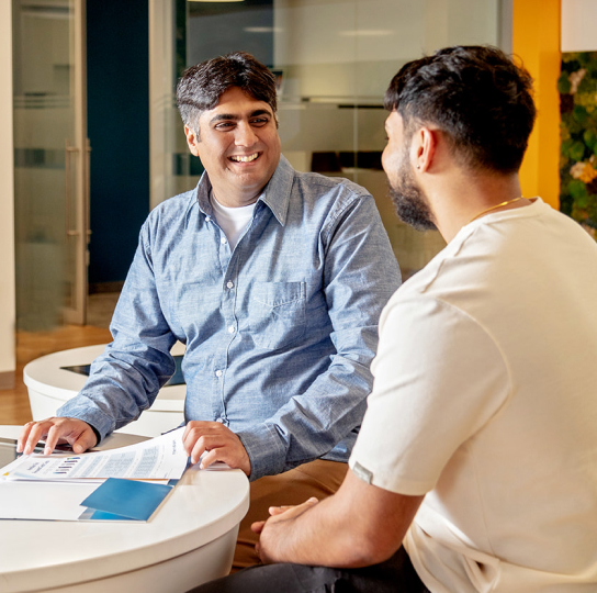 Two brown skinned men sit at a table in a Meridian branch. They are reviewing documents from a folder and talking. 