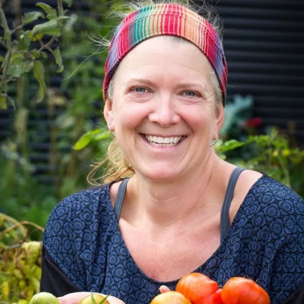 A smiling woman in a garden holding several green and red tomatoes.