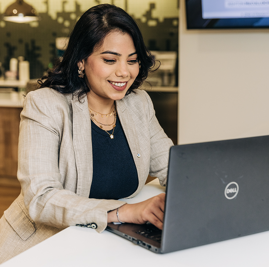 A smiling woman with medium skin tone and dark hair works at a laptop. Meridian employee. Ancaster branch.