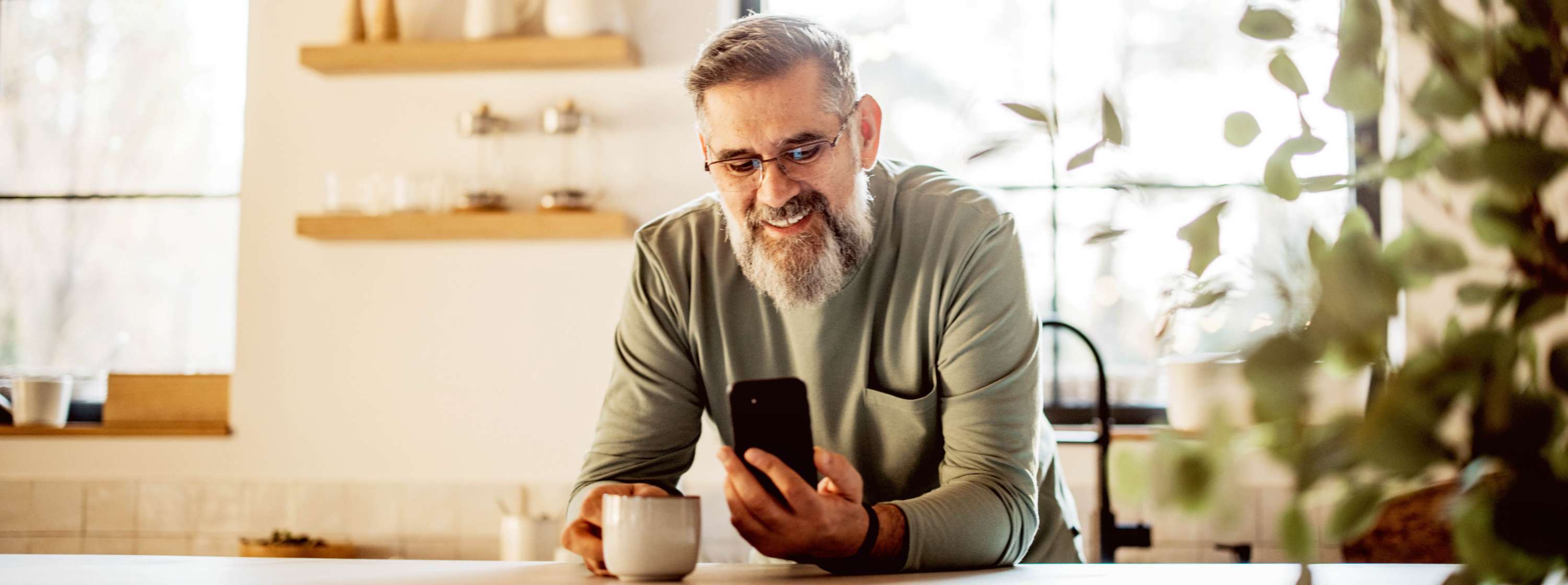 A middle-aged man with grey hair and long scruffy beard, sitting at a table, holding a smartphone in one hand and a cup in the other in a kitchen, with natural light coming through the window behind him.