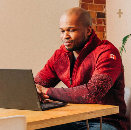 Meridian Member from Racing to Zero, a dark skinned man wearing jeans and a red sweatshirt, sits at a table with his laptop.