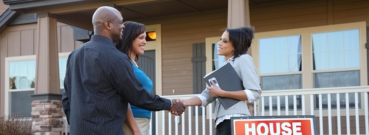 A smiling couple shakes hands with a realtor outside, in front of their new home.
