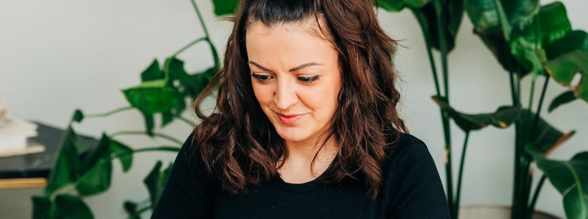 Young woman with medium skin tone and short, dark-brown hair is sitting at a desk looking at her laptop. She is wearing a long sleeved black shirt and there are many potted plants in the background.