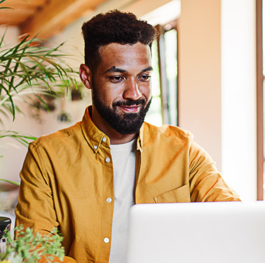 A young black man smiling at his laptop is a plant-filled, sunny room.