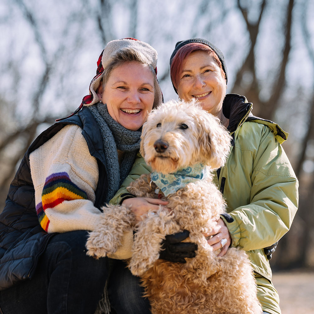 Two women kneeling and hugging their curly-haired dog in a park.