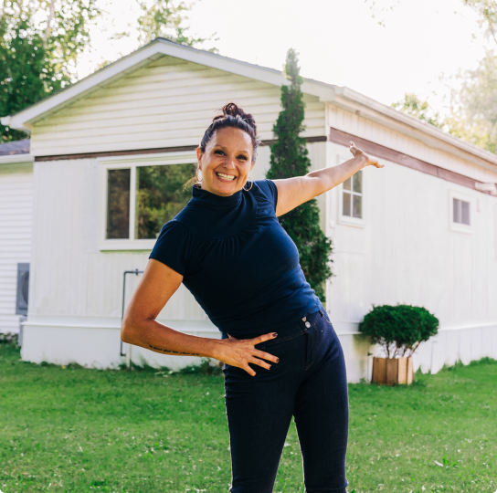 Cher, Meridian Member. She stands in front of a house. Her arm is extended as she gestures toward her new home. 