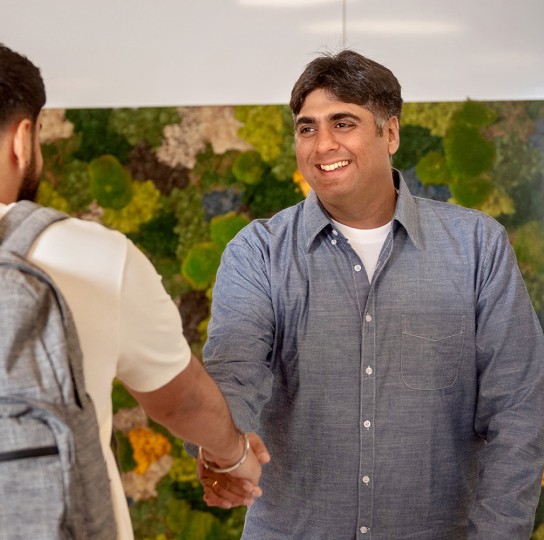 A young man with a backpack shakes hands with a smiling older man with medium tone skin.