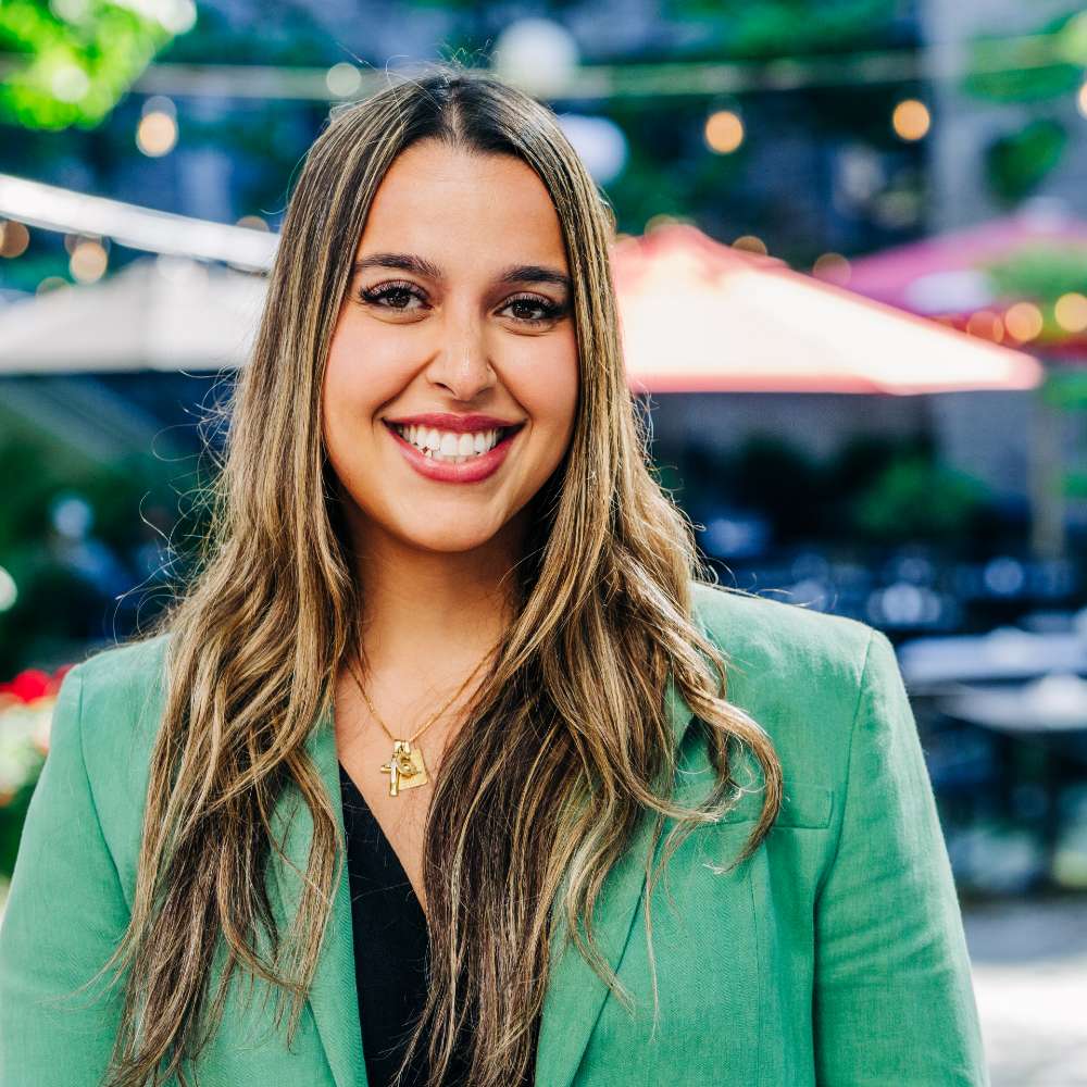 Young woman with medium skin tone and highlighted blonde, long hair is standing outside in front of a restaurant patio. She is wearing a green blazer