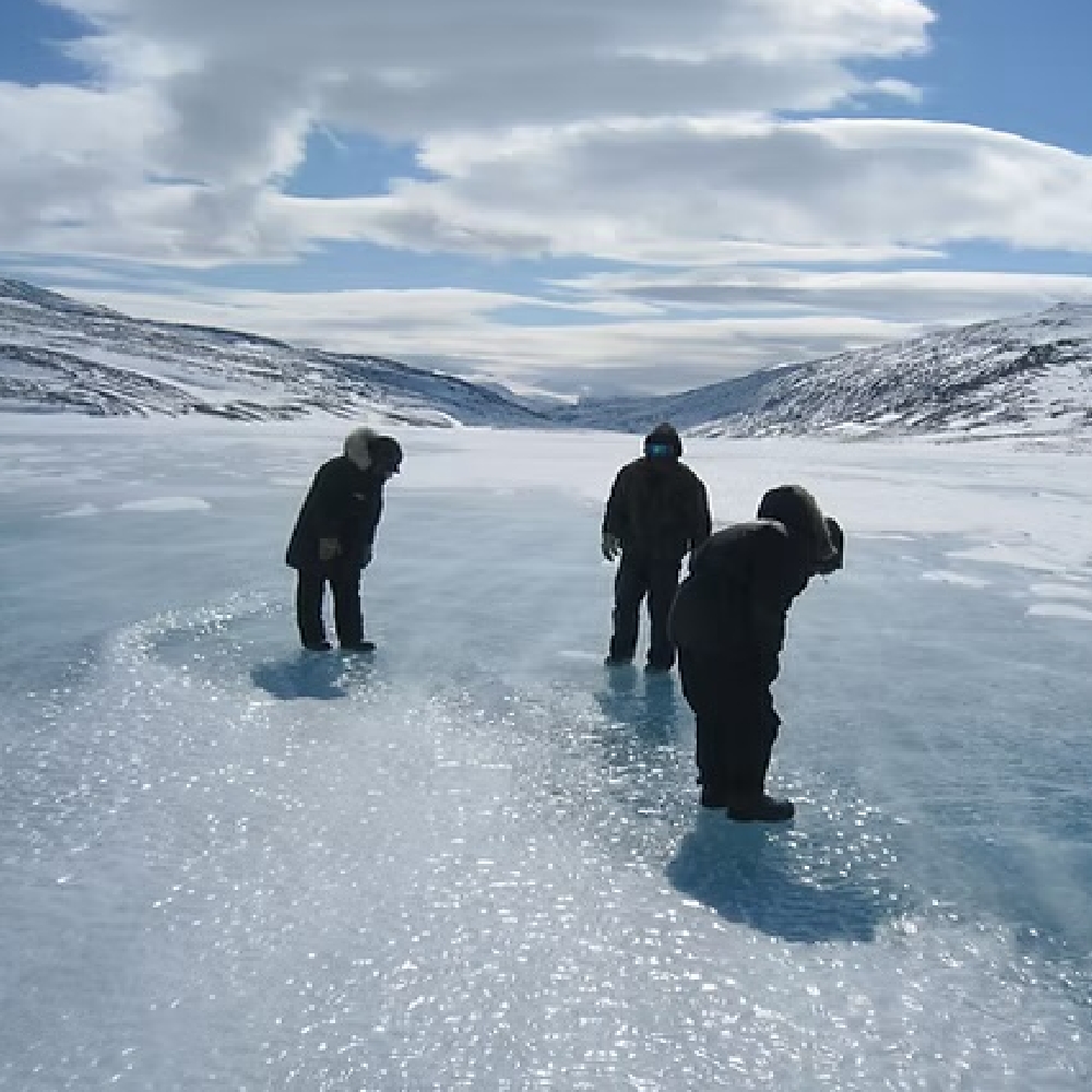 Three people in parkas standing on a frozen lake looking down at the ice.