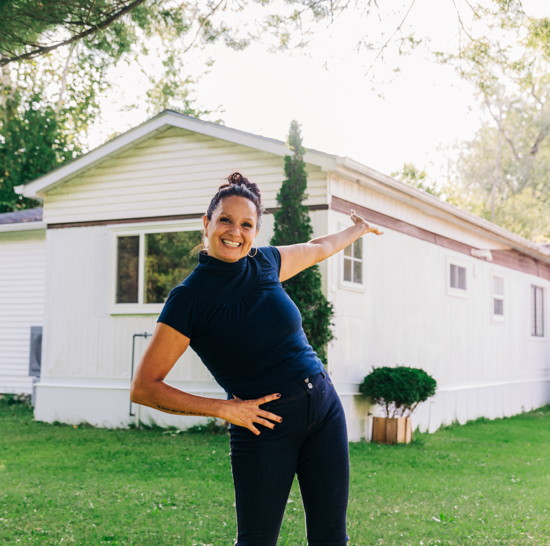 Smiling light skinned woman stands in front of a house, one hand extended toward the property to show it off.